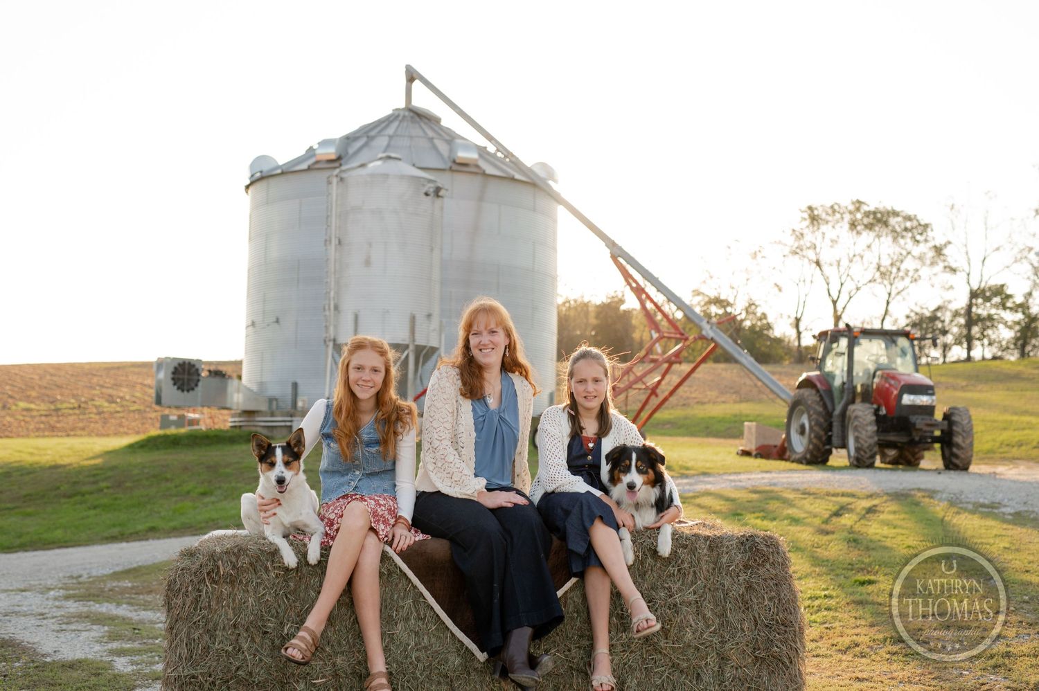 Three people and two dogs sit on hay bale in front of grain silo during sunset.