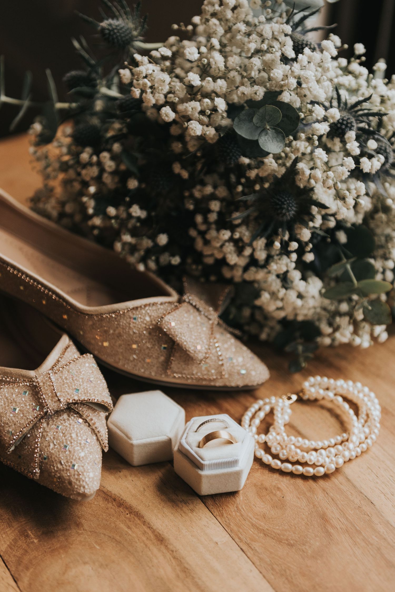 Elegant wedding accessories including sparkly shoes, pearl necklace, ring boxes and baby's breath flowers on wooden surface.