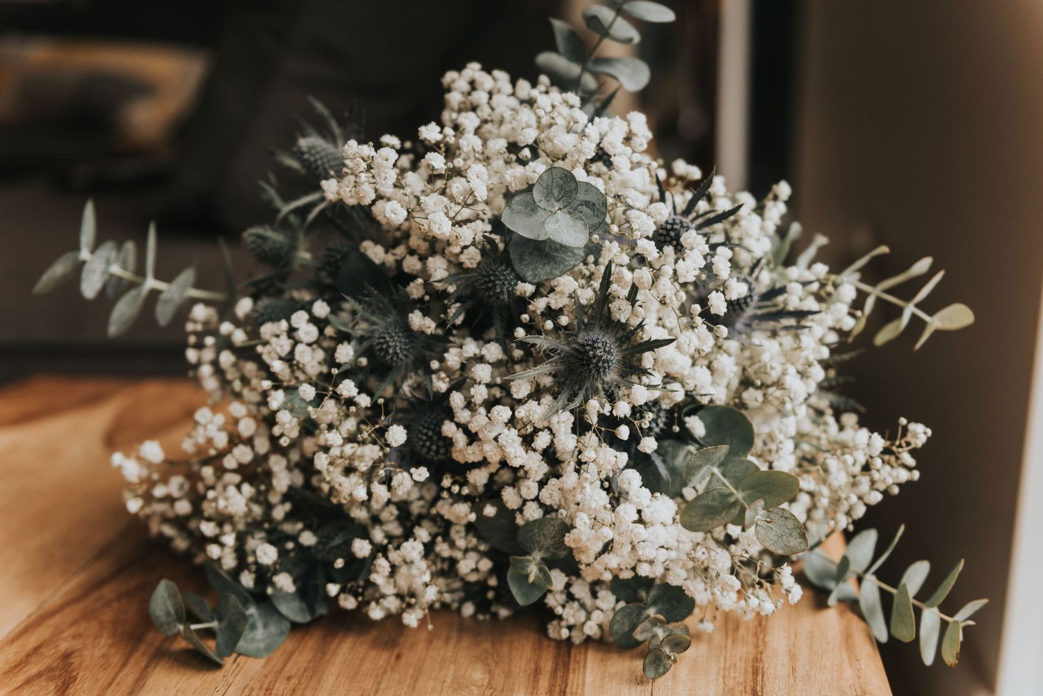 A rustic floral arrangement with white baby's breath and eucalyptus leaves sits on a wooden surface.