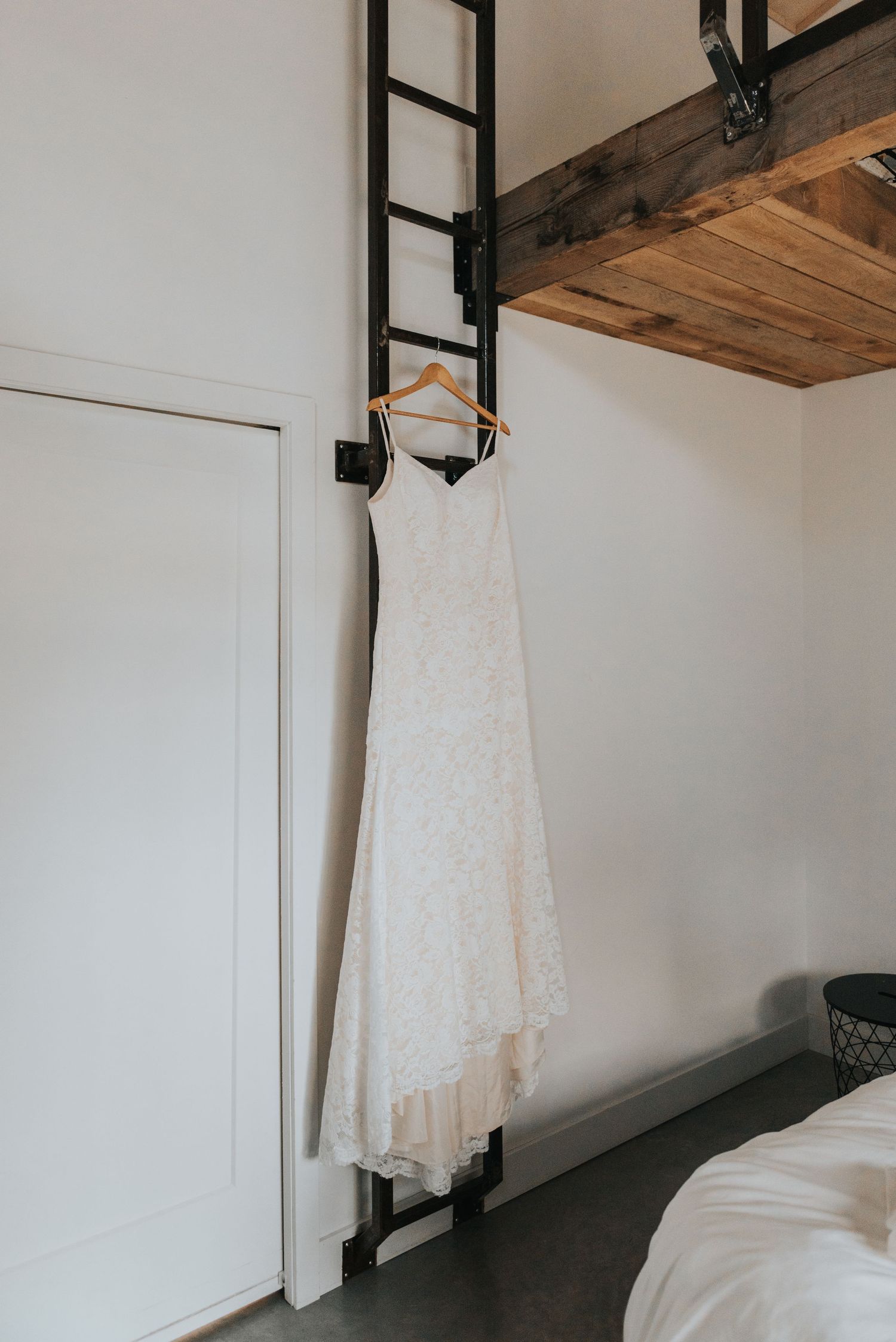 A white wedding dress hangs from a wooden ladder against a minimalist white wall with rustic wooden beams.