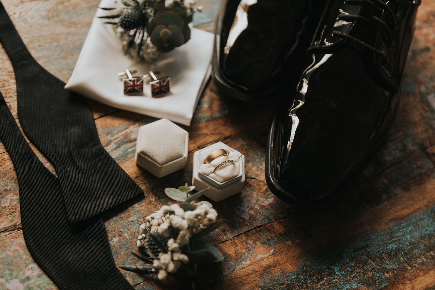 Wedding accessories including black dress shoes, socks, cufflinks and small floral decorations on a rustic wooden surface.