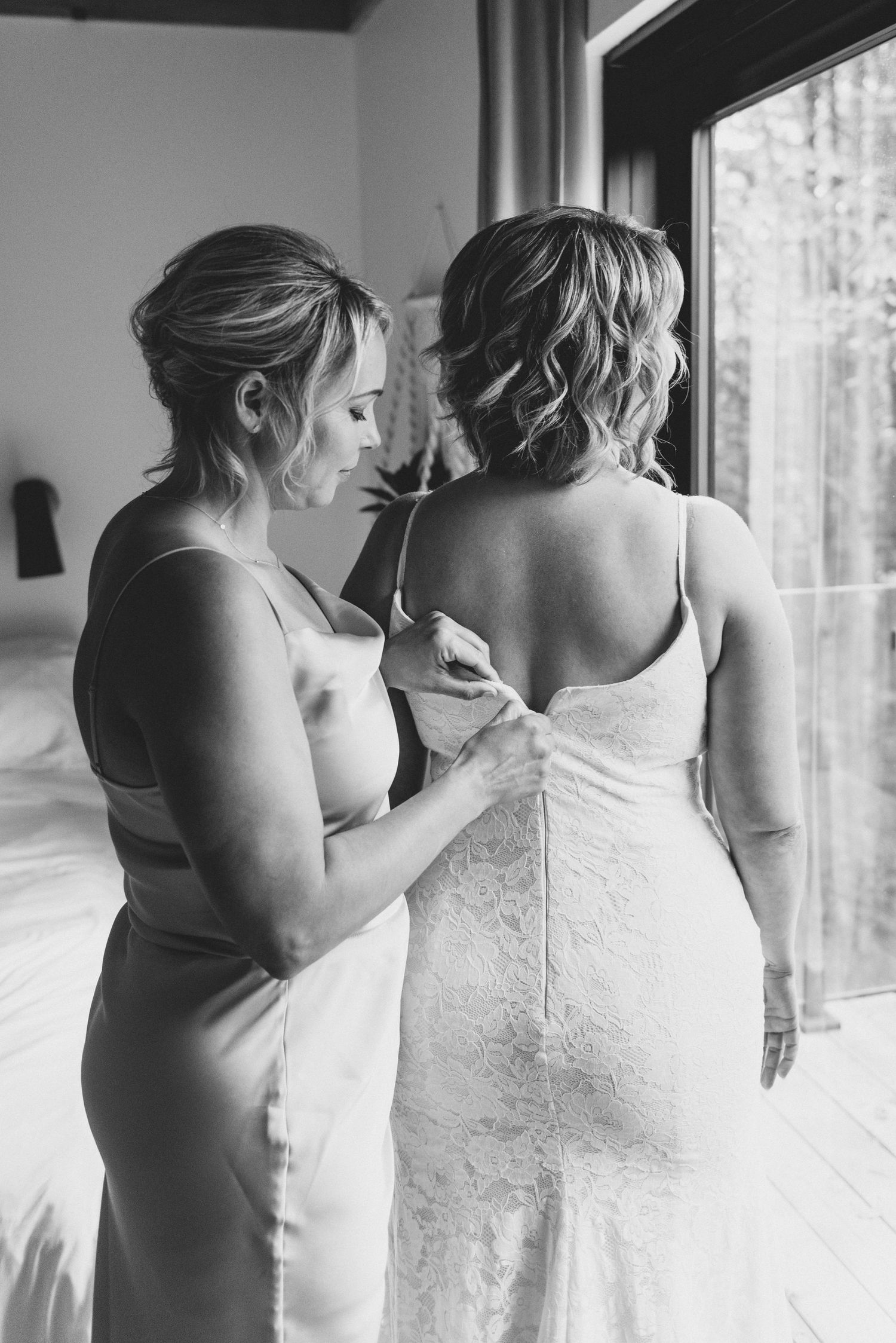 A tender black and white moment during wedding preparations as a woman helps adjust another's elegant lace wedding gown.