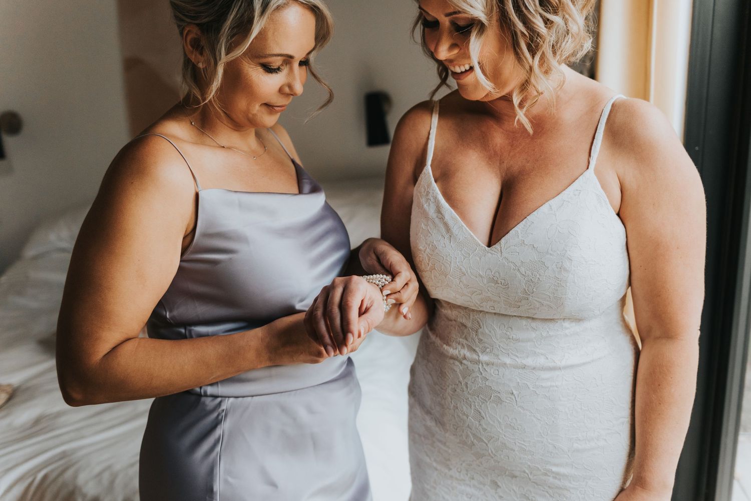 Two women share a tender moment while wearing elegant satin dresses in a softly lit indoor setting.