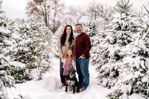 A family poses with their black dog among snow-covered evergreen trees in a winter wonderland setting.
