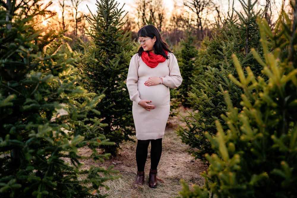 Expectant mother wearing a beige sweater dress and red scarf stands among evergreen trees at sunset.