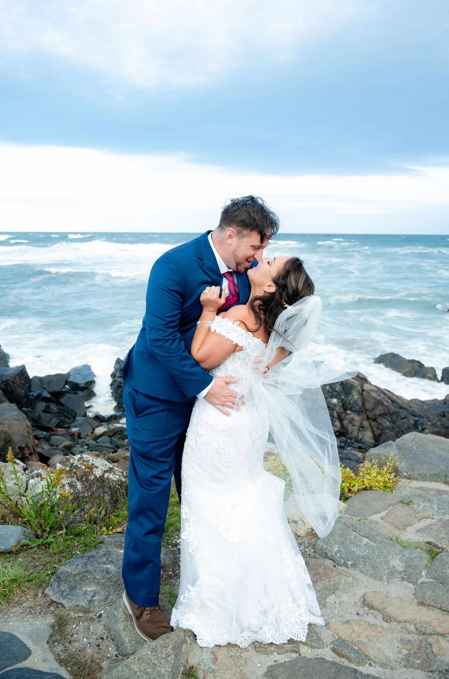 Romantic coastal wedding photos captured on rocky cliffs with waves crashing in the background.