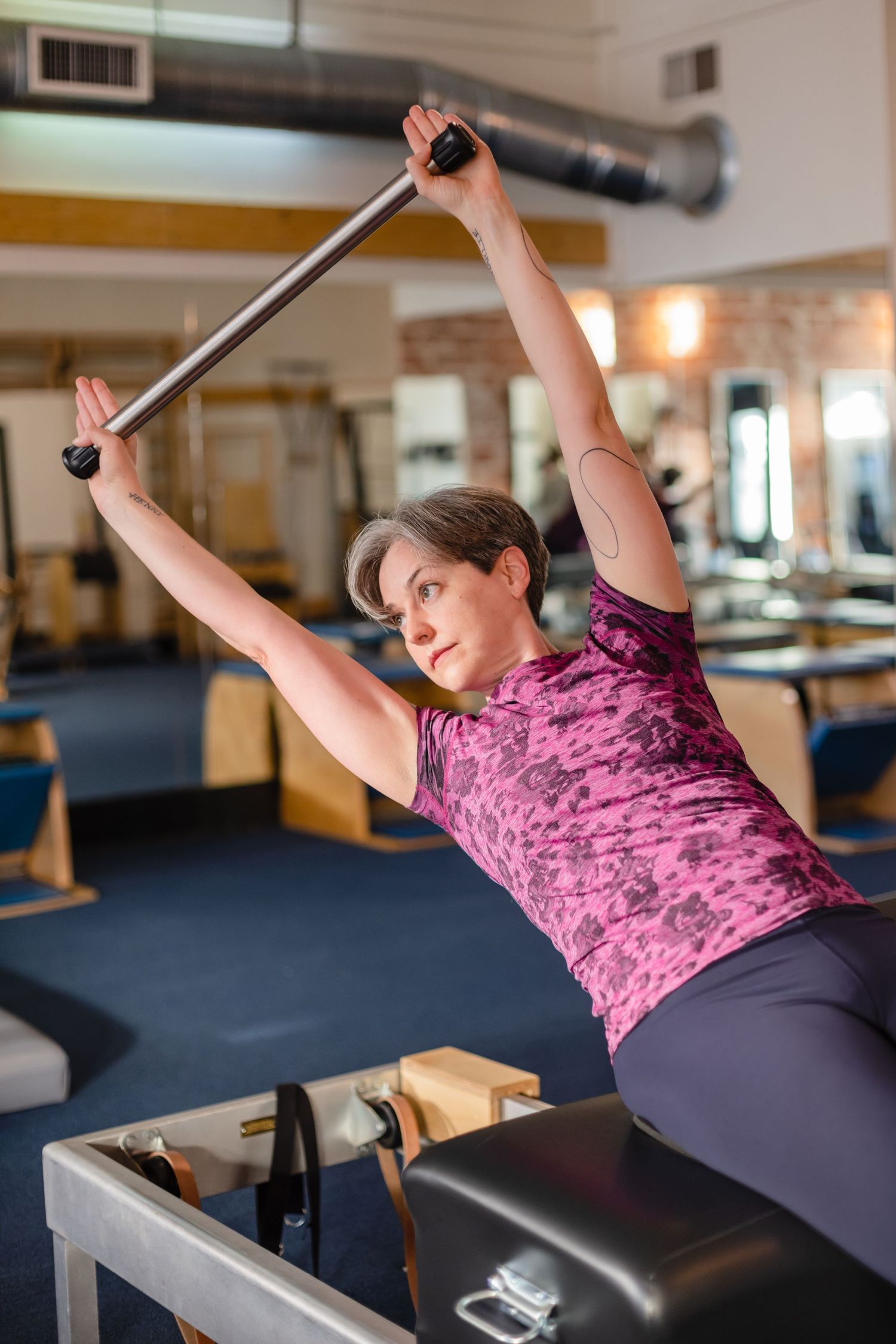 Person exercising on reformer Pilates machine wearing pink top and grey leggings in fitness studio.