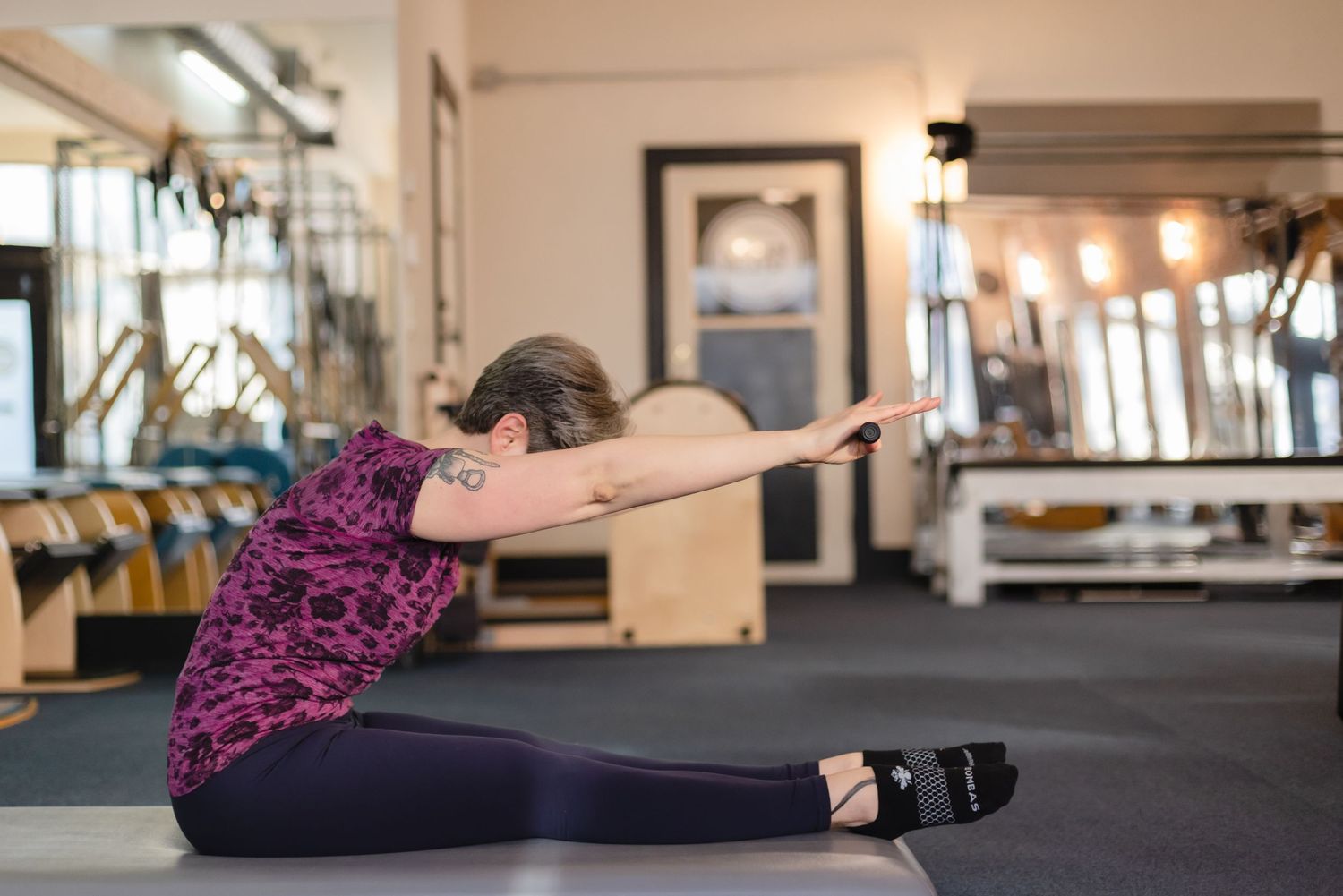 Person performing a seated forward fold stretch on a black mat in a fitness studio with exercise equipment.