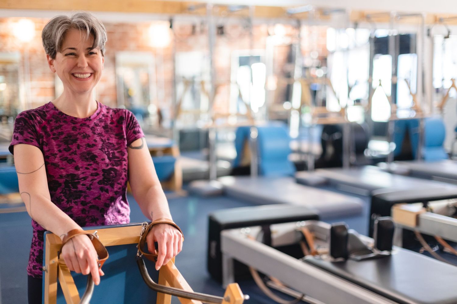 Pilates instructor in burgundy floral dress stands by exercise equipment in a bright fitness studio.