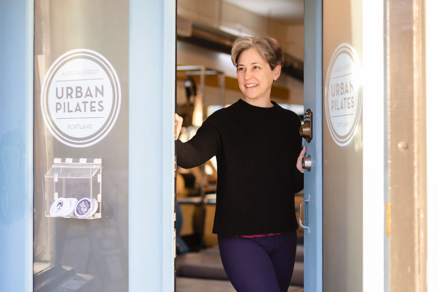 Someone stands in the doorway of Urban Pilates studio smiling while leaning against the glass entrance.