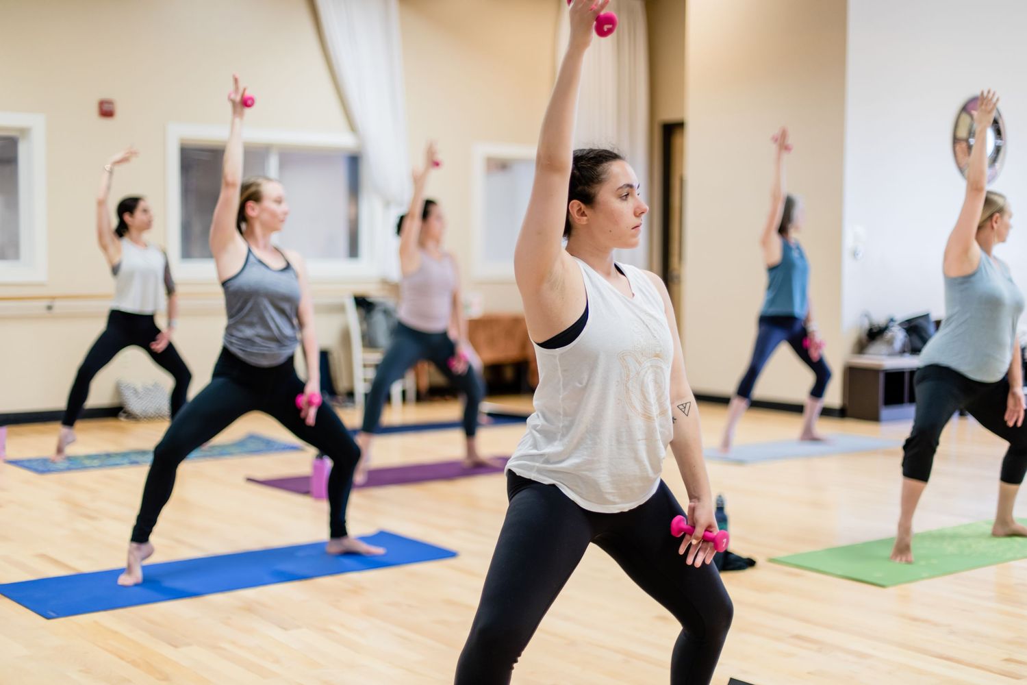 Fitness class participants exercising with dumbbells in warrior pose on colorful yoga mats at a bright studio.