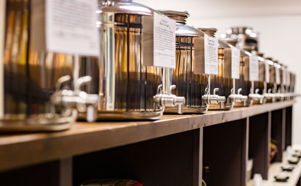 Long row of stainless steel olive oil dispensers on wooden shelving.