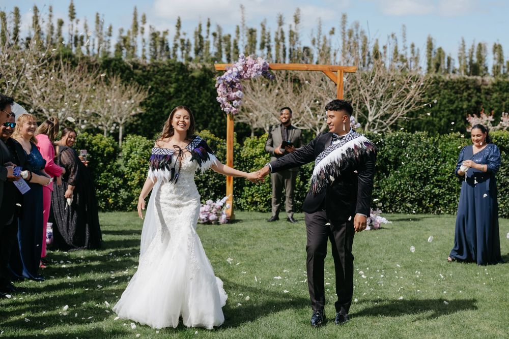 Bride and groom celebrate with wedding guests during an outdoor garden ceremony as flower petals fall around them.