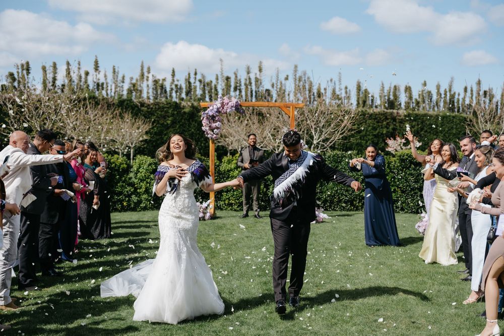 Wedding guests celebrate by throwing confetti in a sunny garden setting with tall ornamental grass in the background.