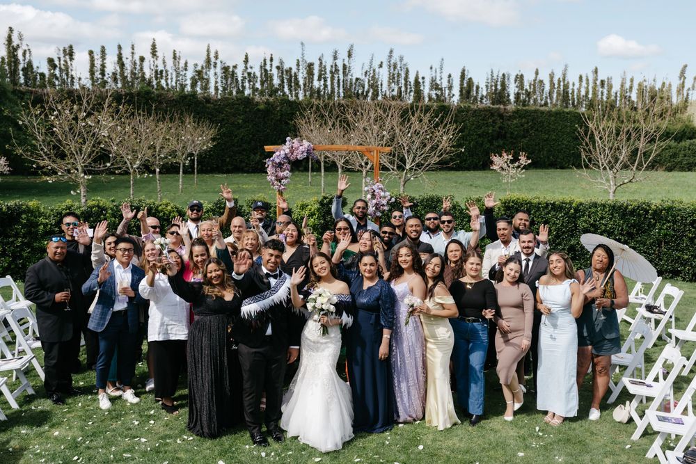 A large group of wedding guests gathered outdoors in formal attire for a group photo in a manicured garden setting.