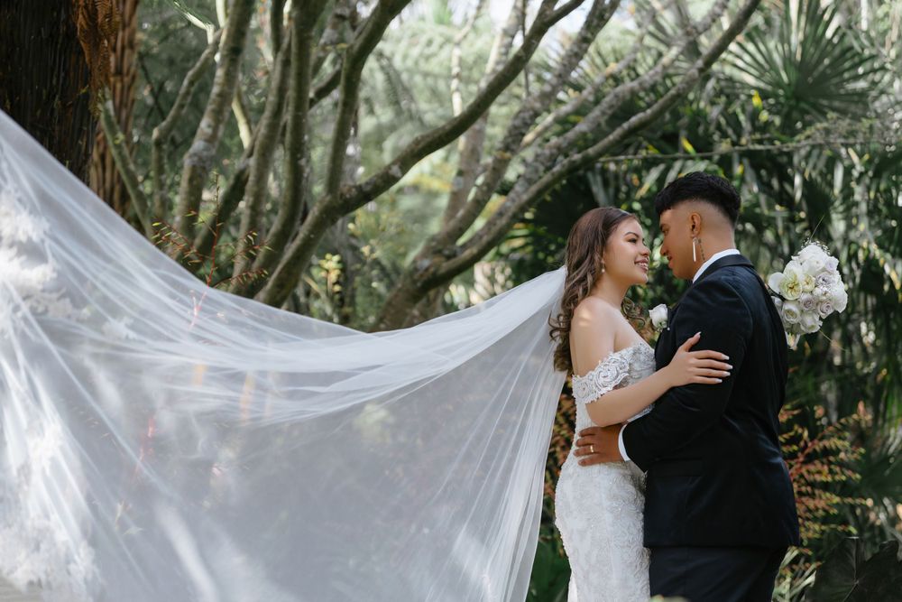 A couple in wedding attire share an intimate moment in a lush garden setting while a white veil flows in the breeze.
