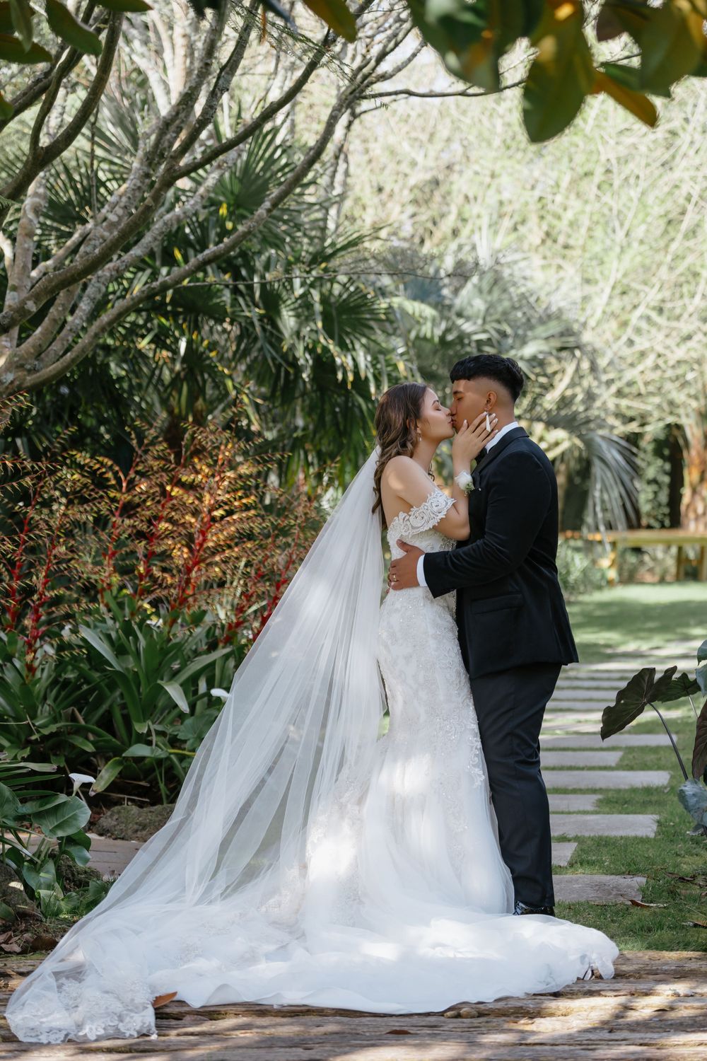 A romantic garden wedding scene with a couple kissing on a stone path surrounded by lush tropical foliage.