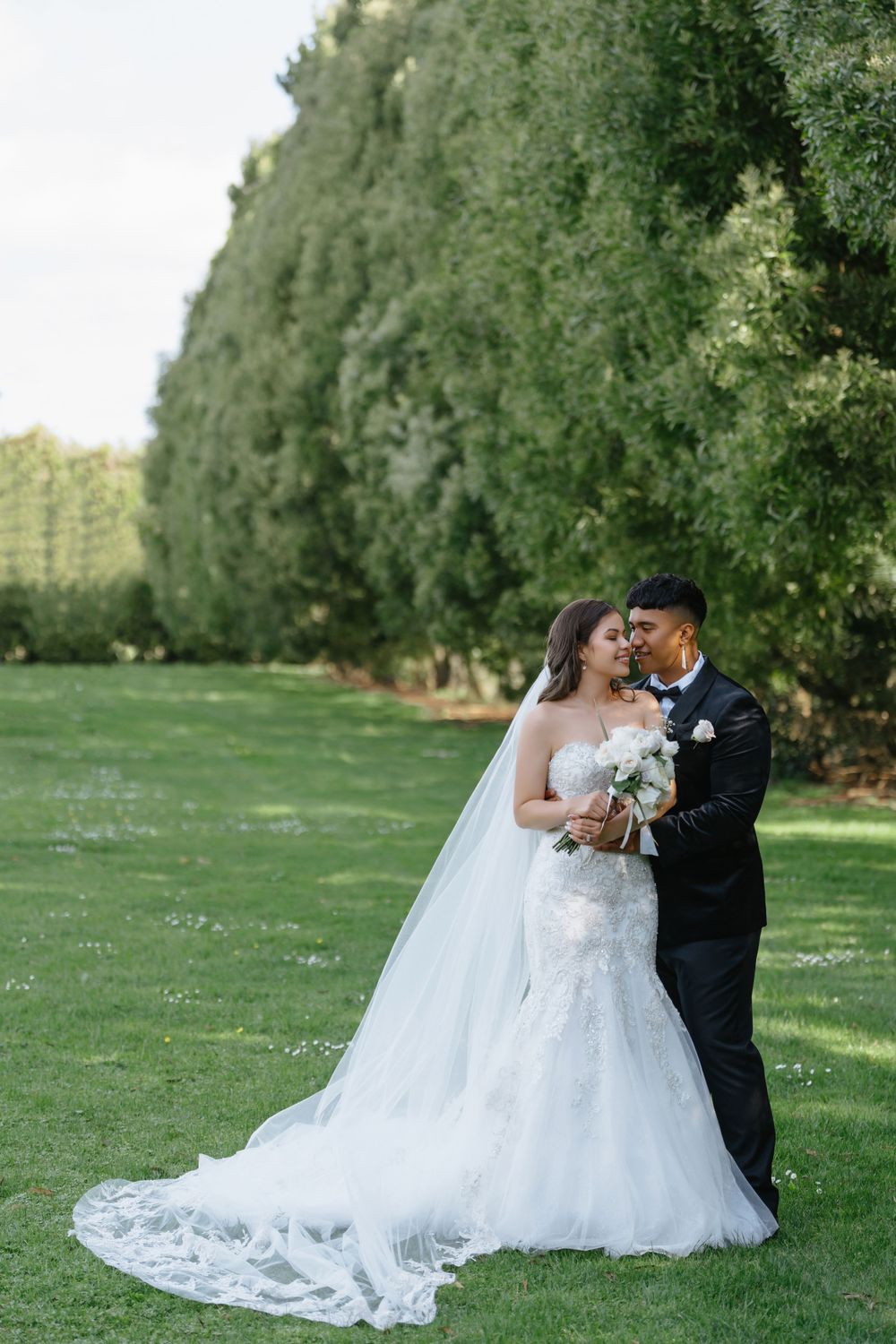 A beautiful wedding couple embraces on a lush green lawn with tall trees creating a natural backdrop on their special day.