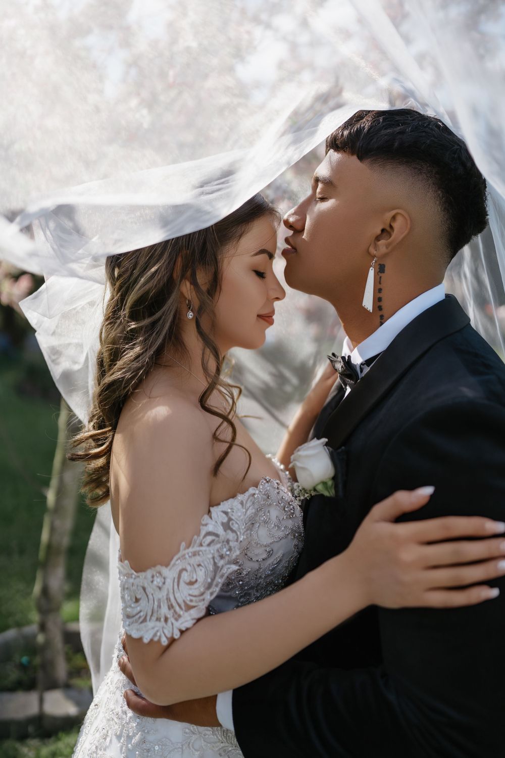A romantic wedding portrait showing a couple embracing under a flowing veil outdoors.