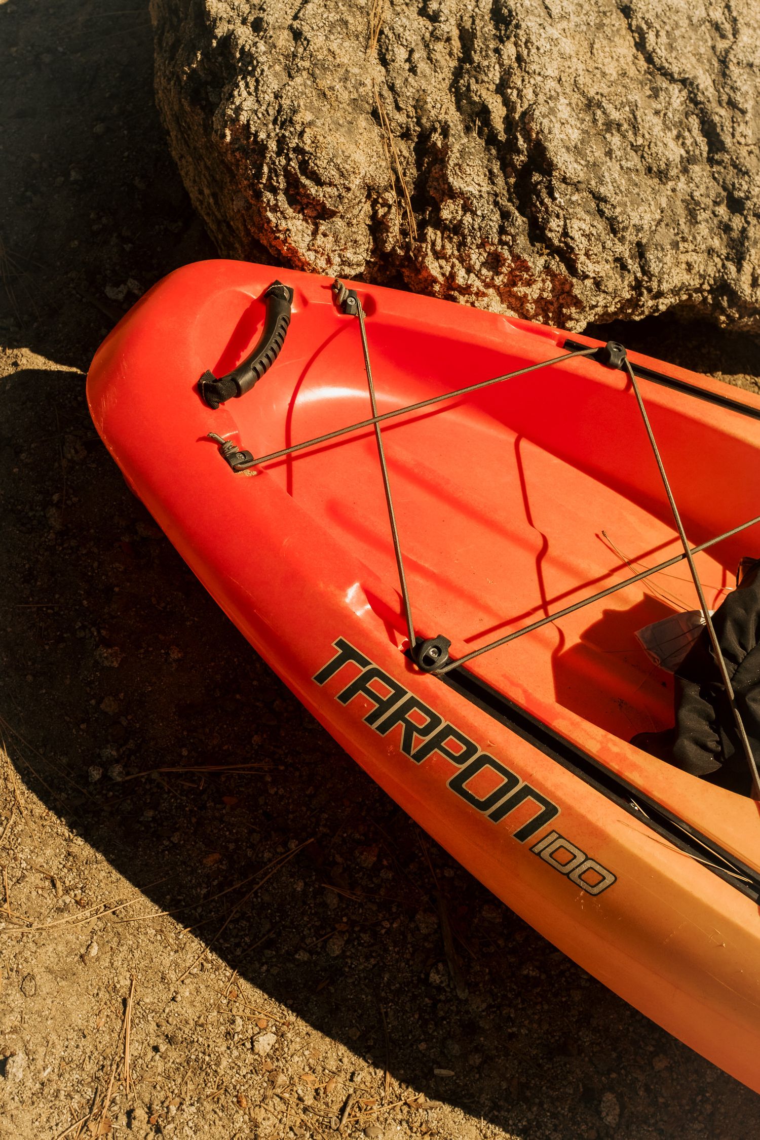 Close-up view of a bright orange Tarpon kayak bow with bungee cord rigging against rocky terrain.
