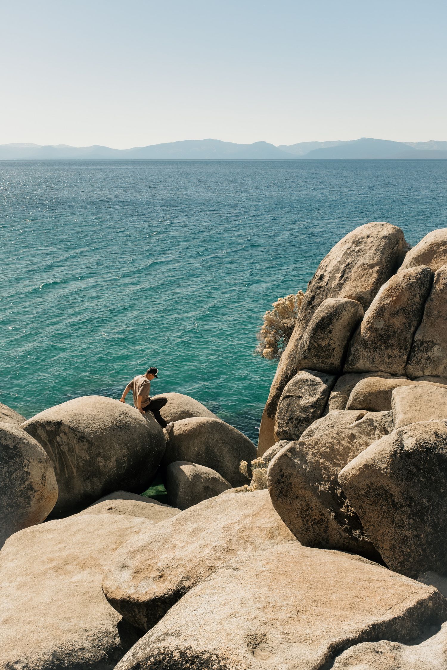 Person sits on large granite boulders overlooking turquoise Lake Tahoe waters with distant mountains in the background.
