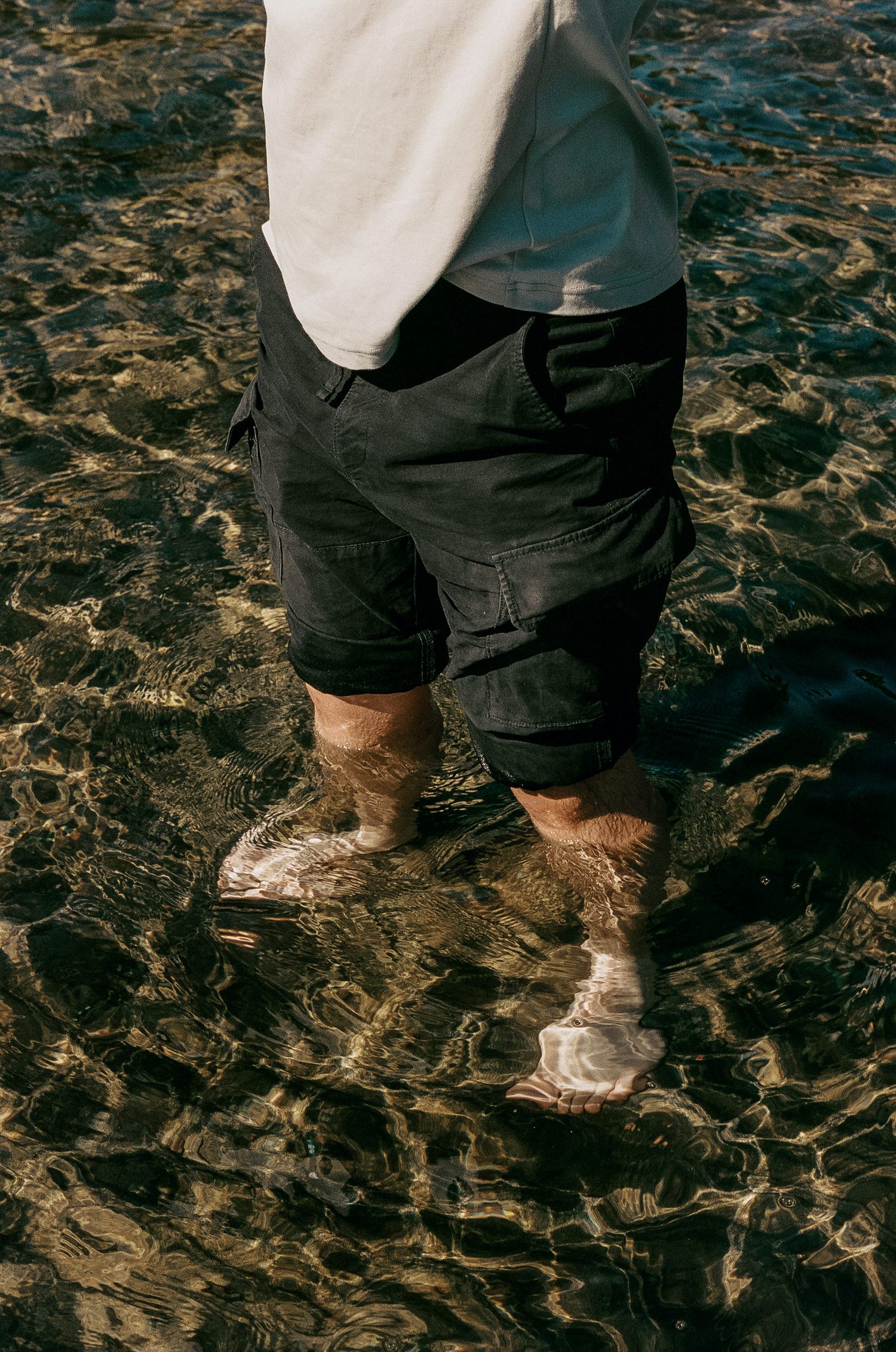 Bare feet standing in shallow dark water wearing black cargo shorts and white shirt captured from waist down.