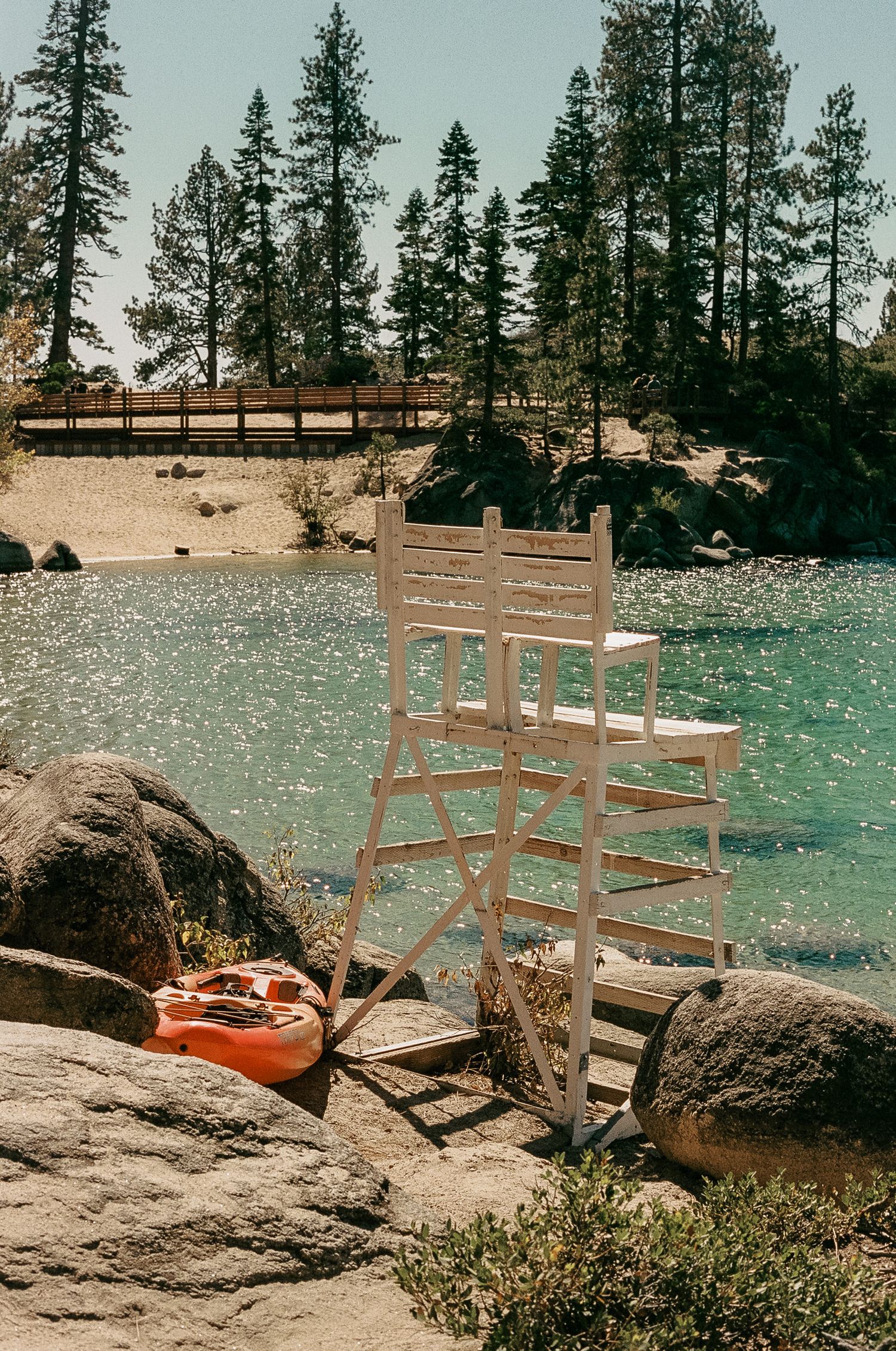 A lifeguard chair sits on rocky shore beside turquoise lake waters with pine trees lining the background.
