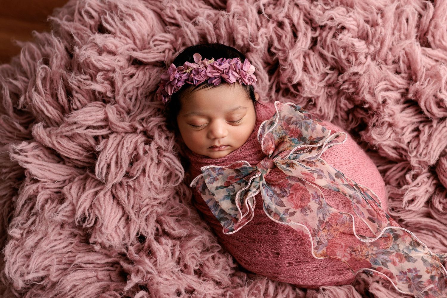 Sleeping newborn wrapped in pink and gray floral swaddle lying on soft mauve fur blanket with matching headband accent.