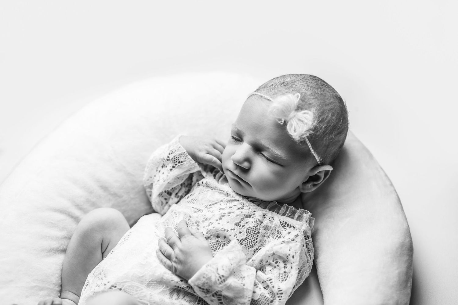 Black and white photo of an infant lying on white bedding wearing a floral dress with a small bow accessory.
