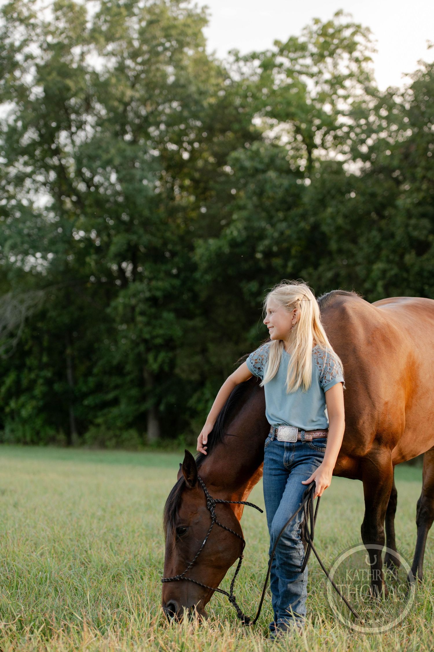 A person in casual attire stands with a grazing horse in a green field at sunset.
