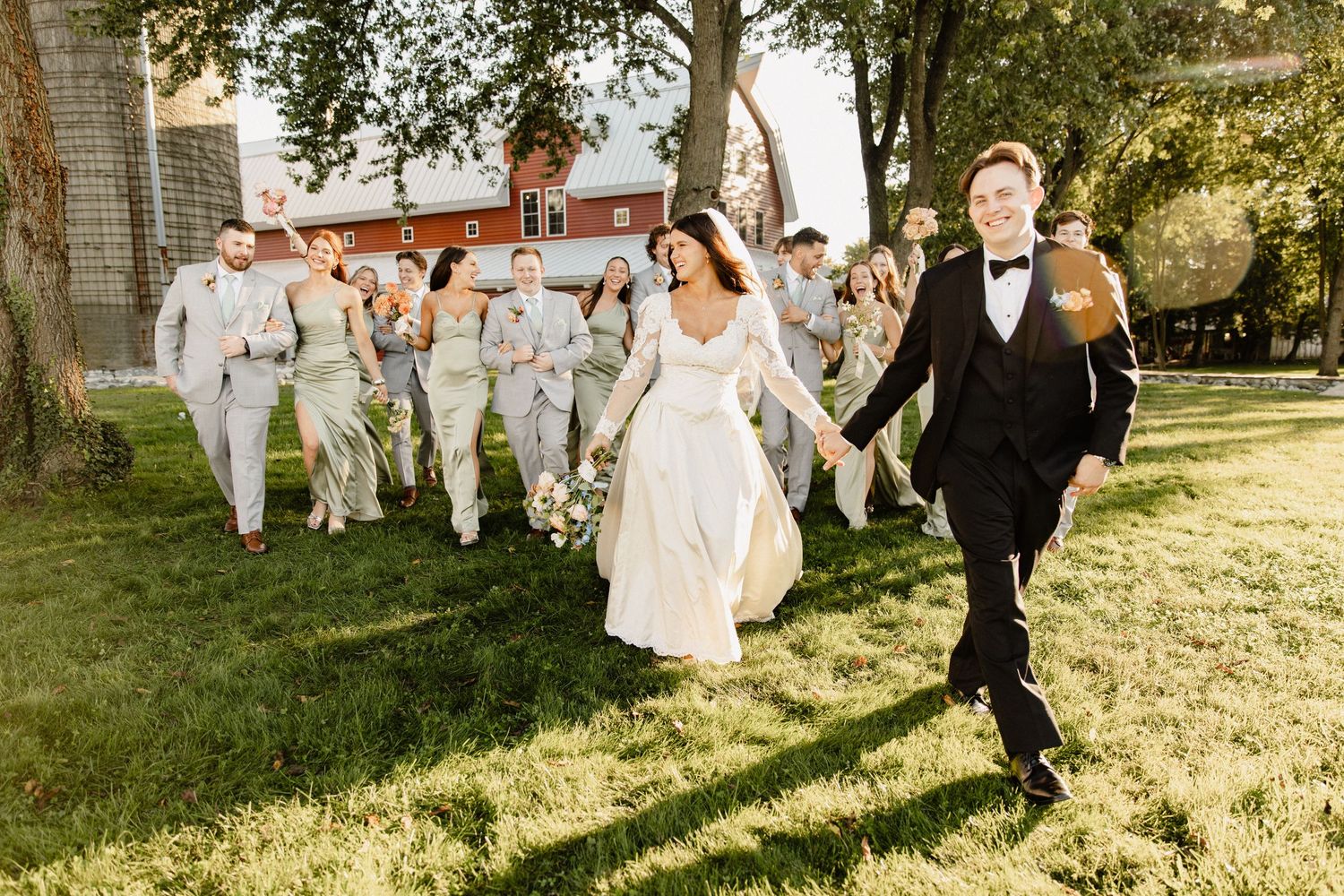 A wedding party walks joyfully across a sunlit lawn with bridesmaids in white dresses.