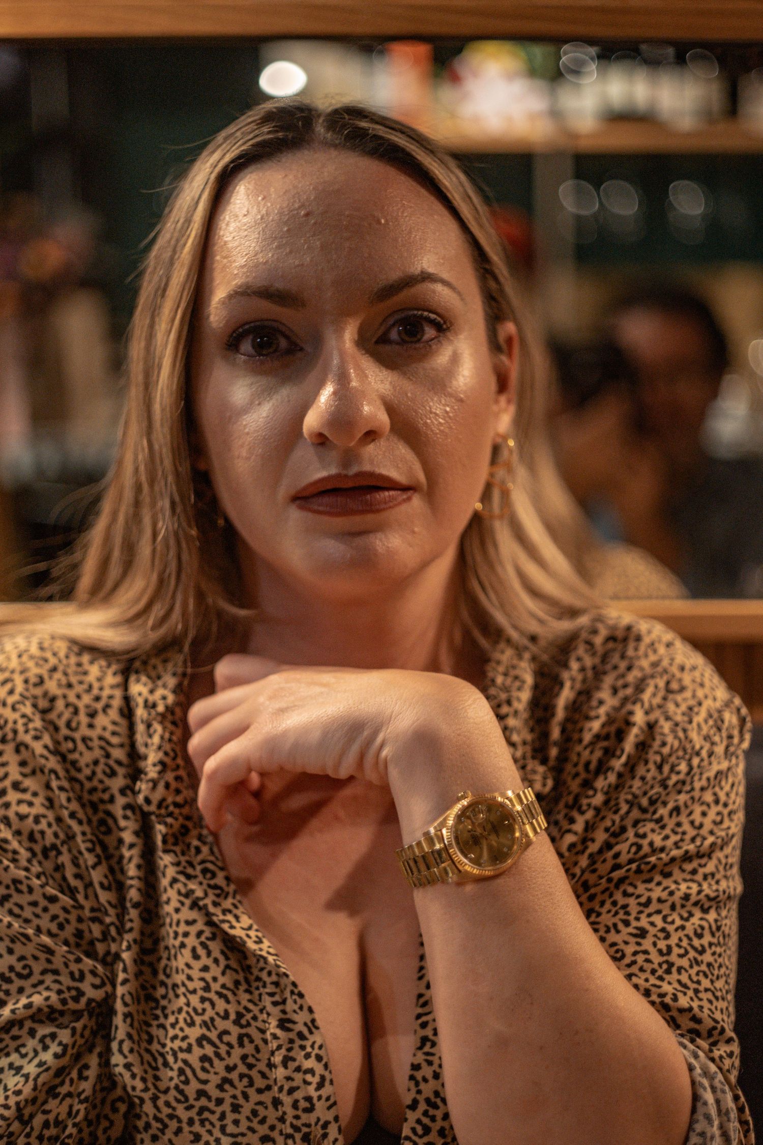 Close-up portrait in warm lighting showing someone wearing a leopard print top and gold watch at a bar.