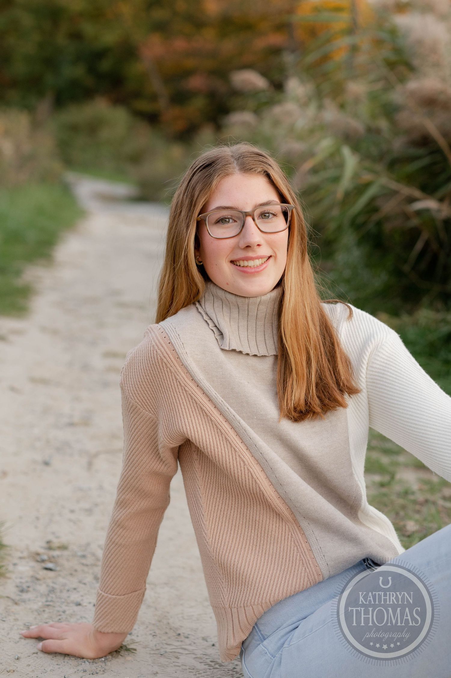 Casual outdoor portrait in beige turtleneck sweater sitting on path with autumn foliage.