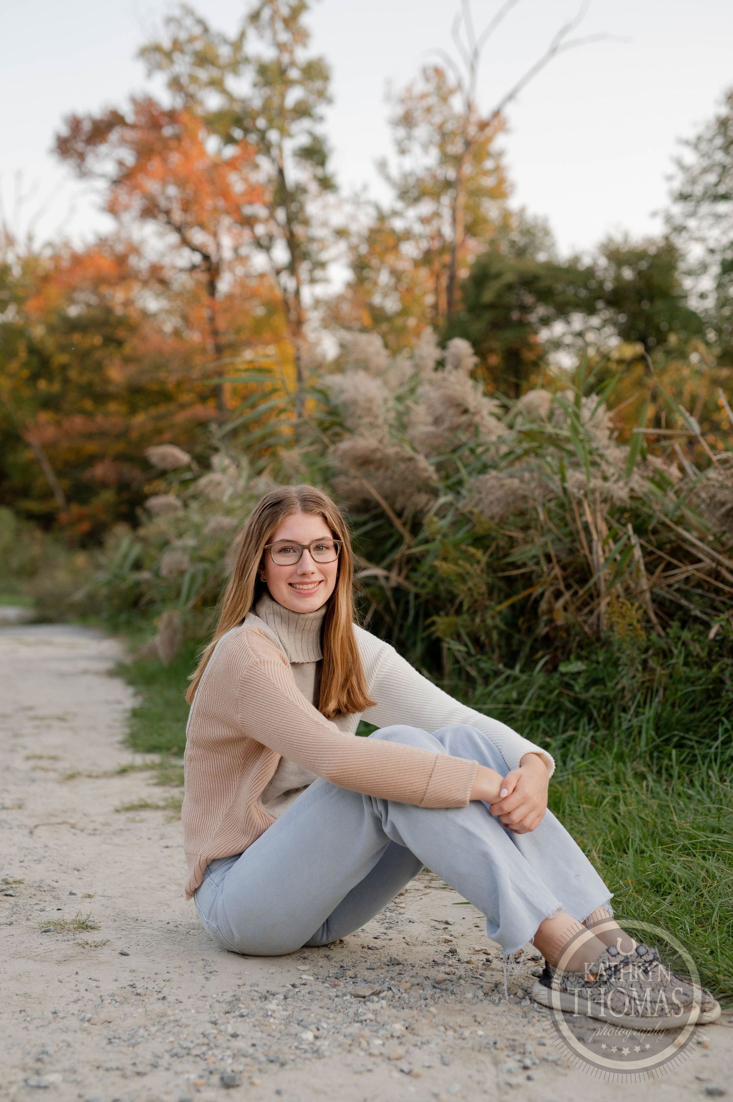 Casual portrait sitting on dirt path with autumn foliage and grasses in background.