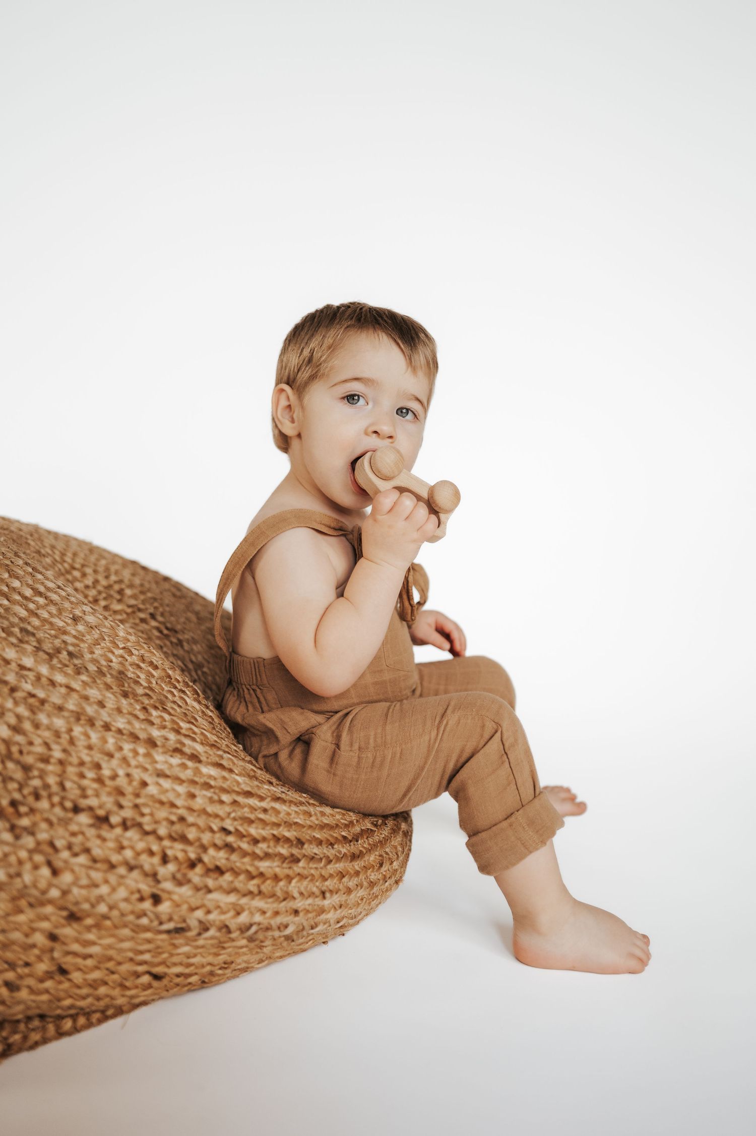 A young child in brown overalls sits on a woven basket while sucking their thumb against a white background.