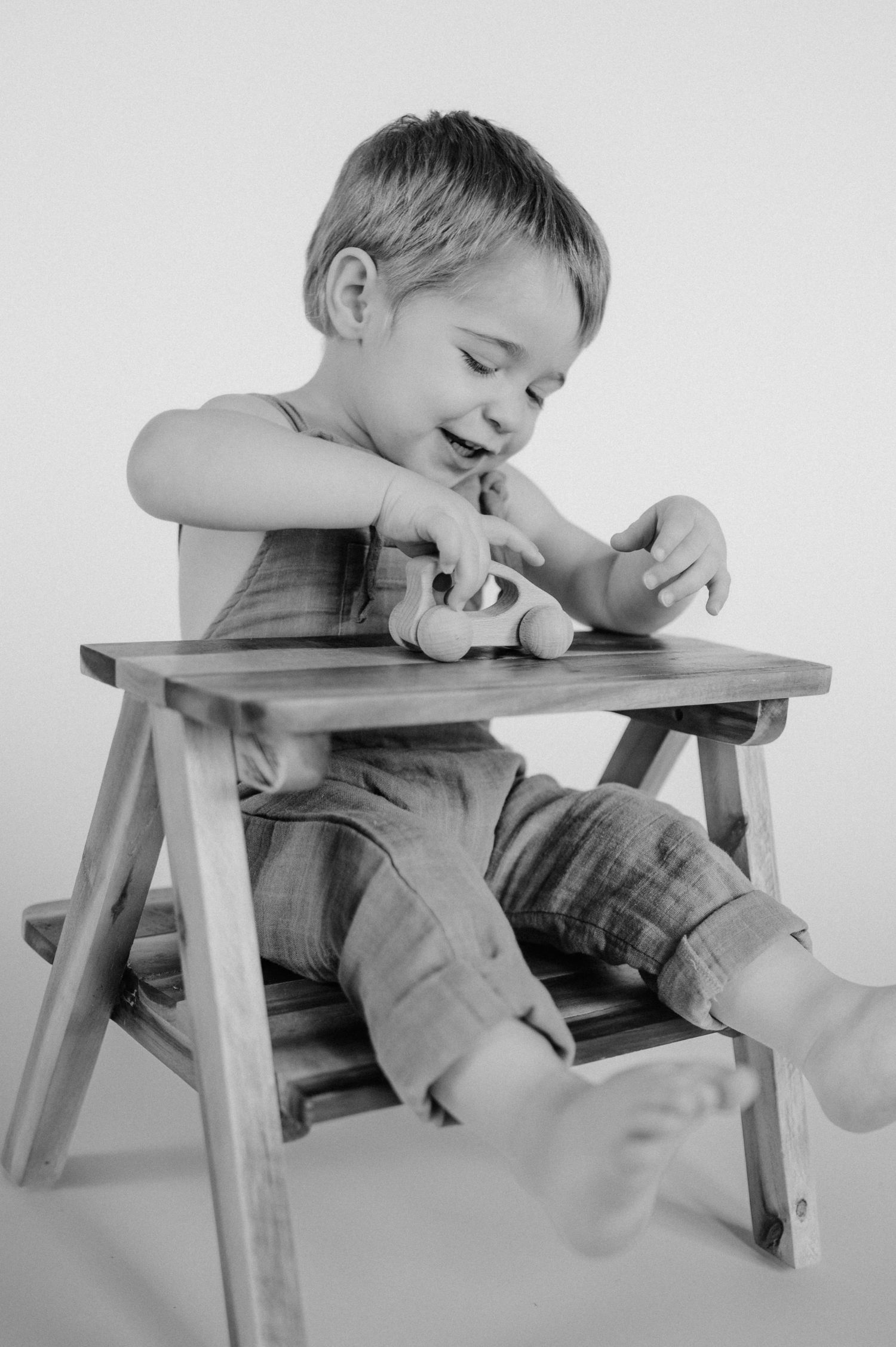 A young child in overalls sits on a wooden chair playing with a toy in a black and white photograph.