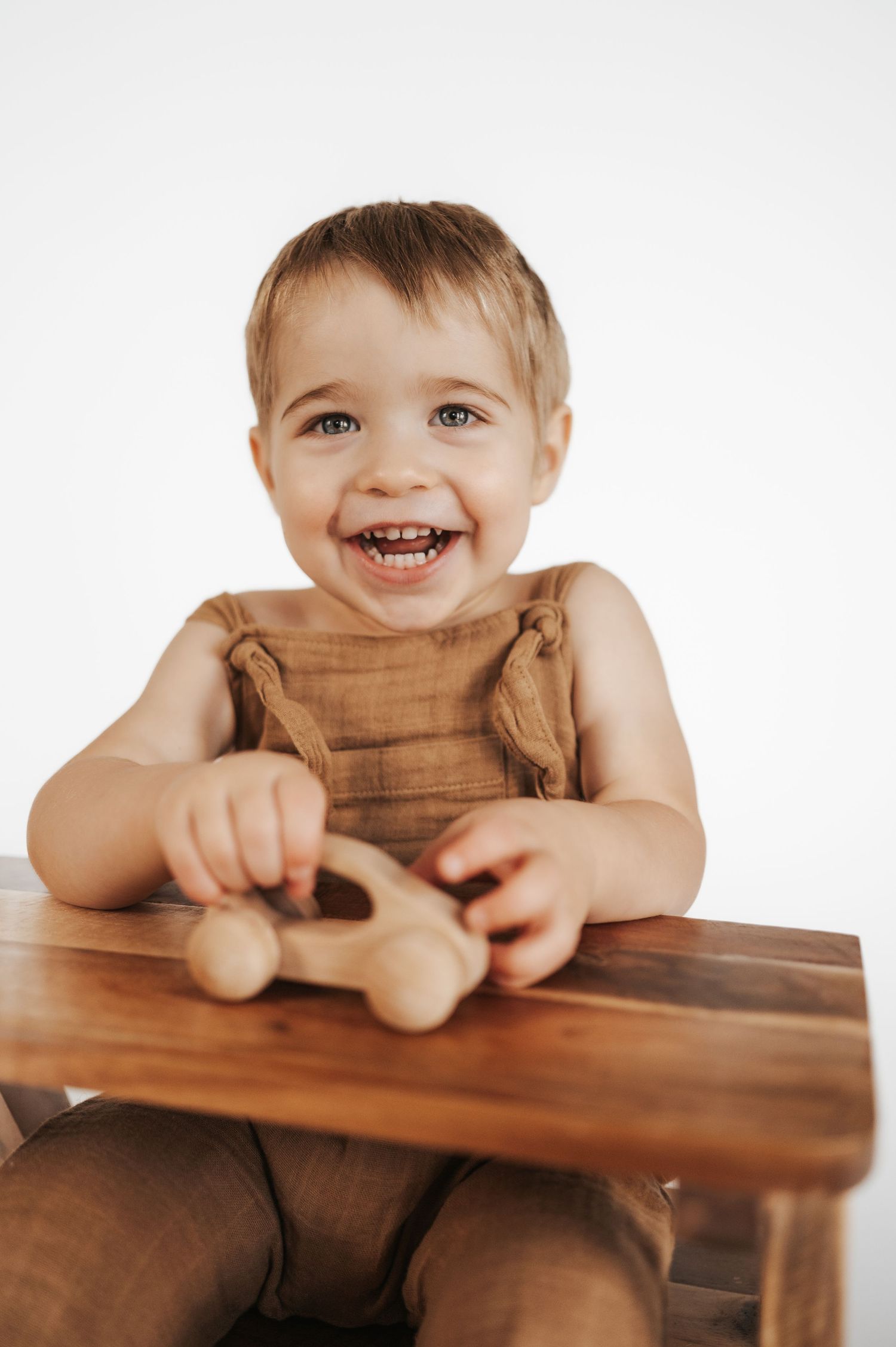 A happy child plays with a wooden toy car while sitting at a wooden table wearing brown overalls.