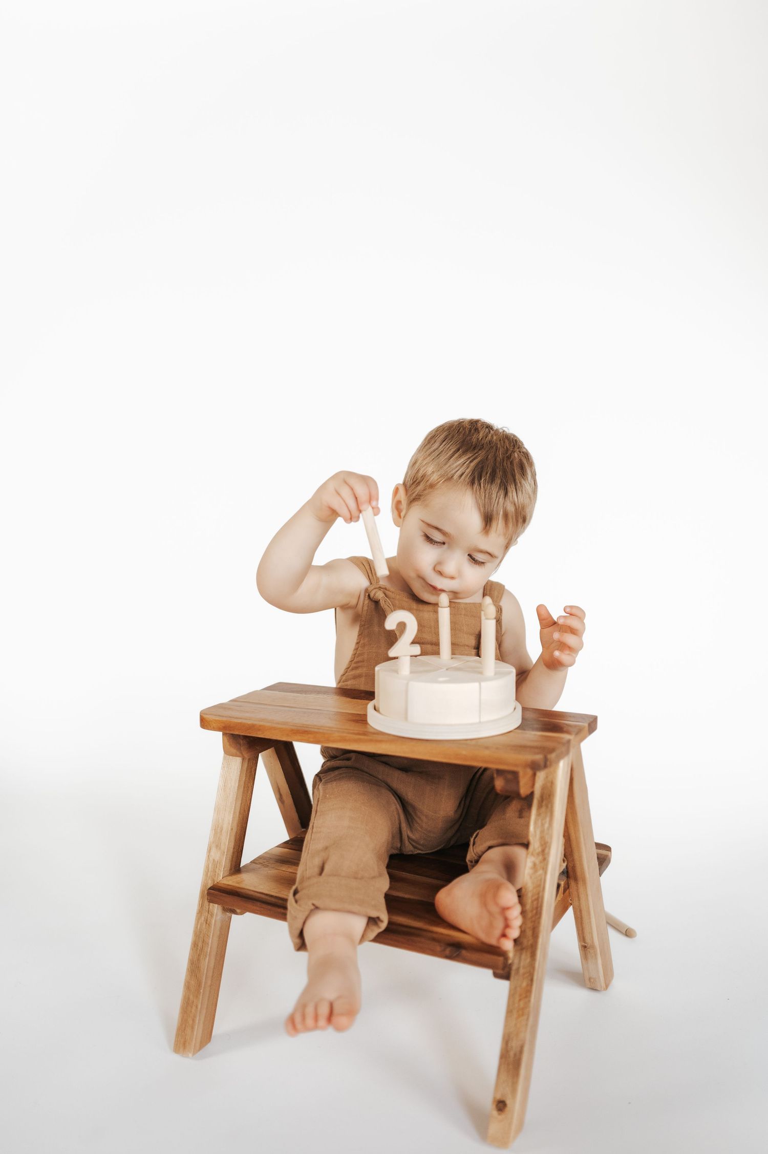 Small child in brown overalls sits in wooden high chair reaching for birthday cake with candles on white background.