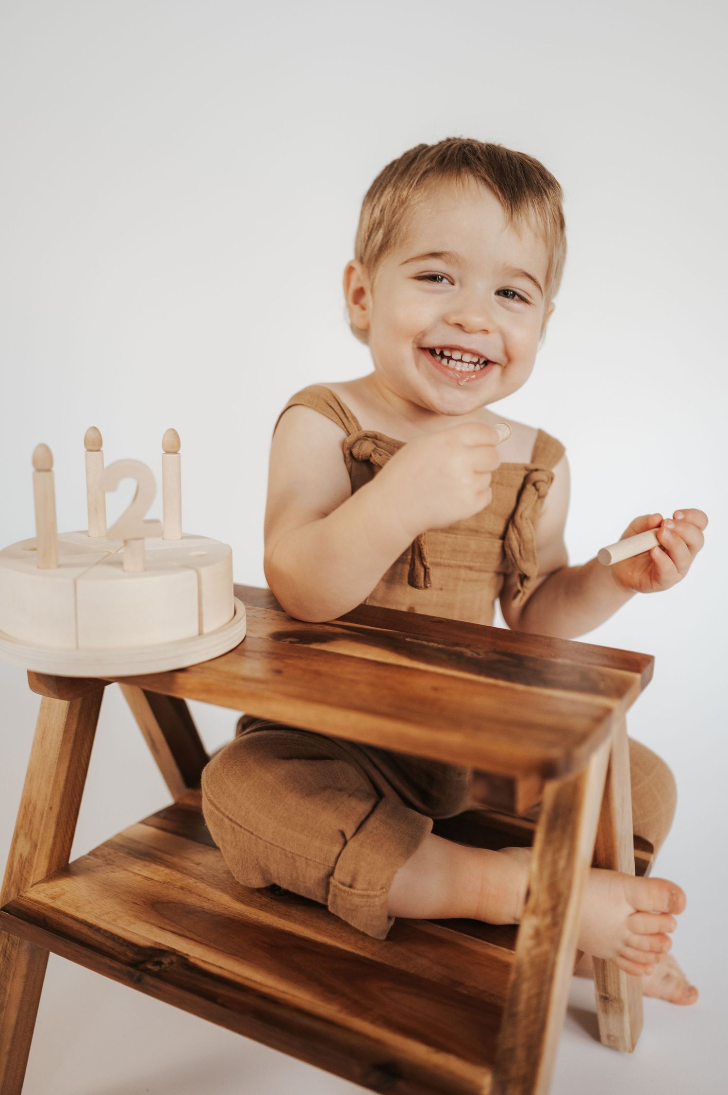 A happy toddler in brown overalls sits at a wooden table with a birthday cake featuring wooden candles.