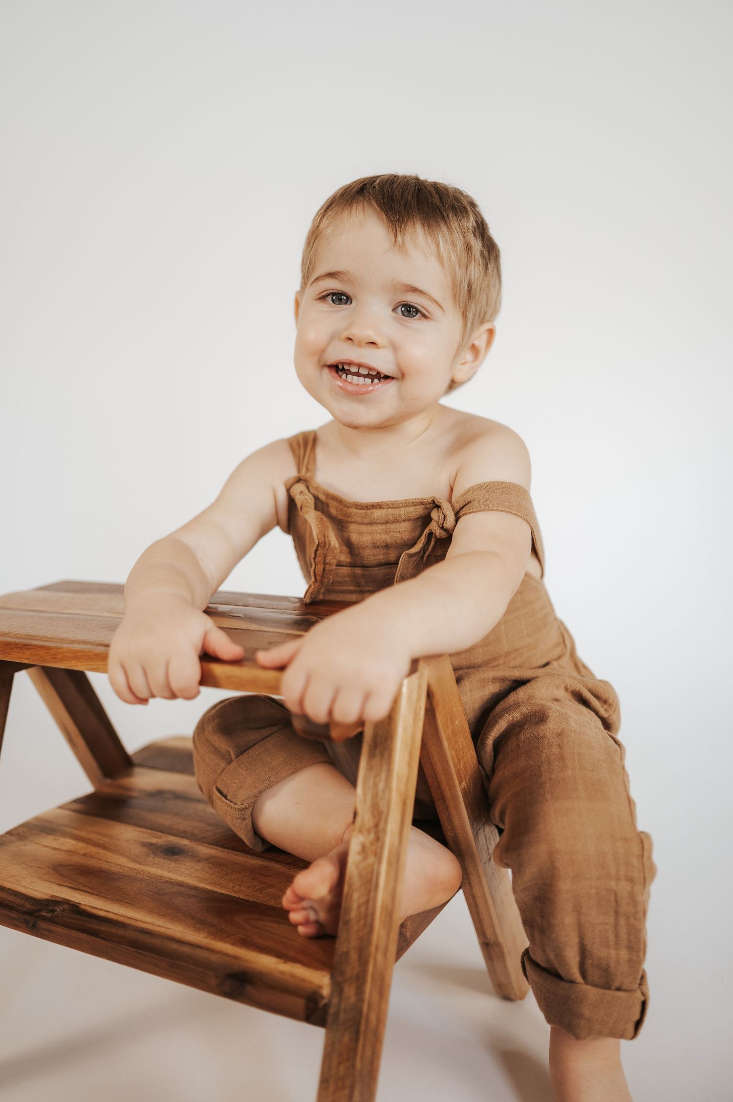 Young child sits on a wooden step stool wearing brown overalls with bare feet dangling.