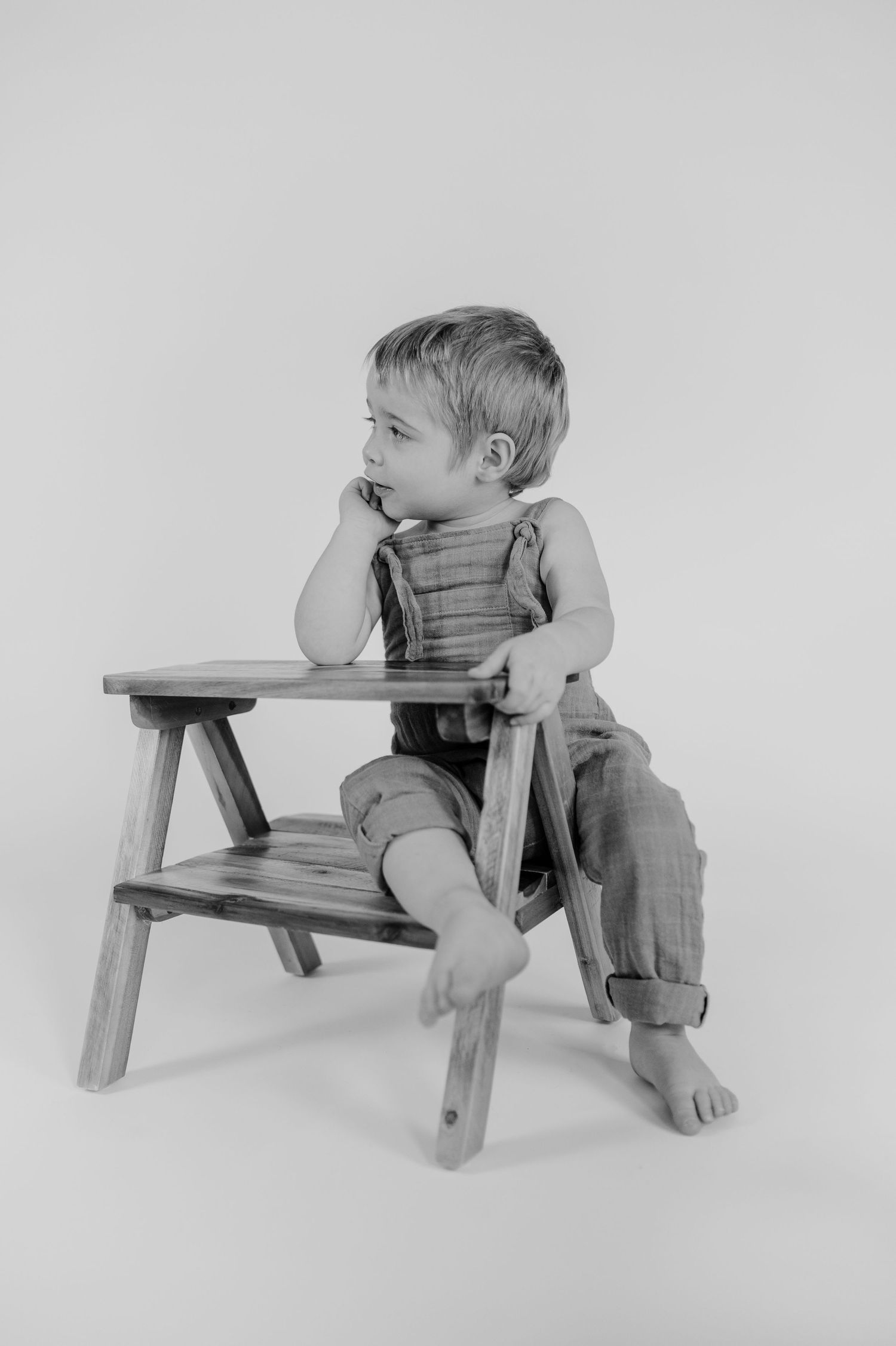 Black and white photo showing a small child in overalls sitting on a wooden stool looking thoughtfully to the side.