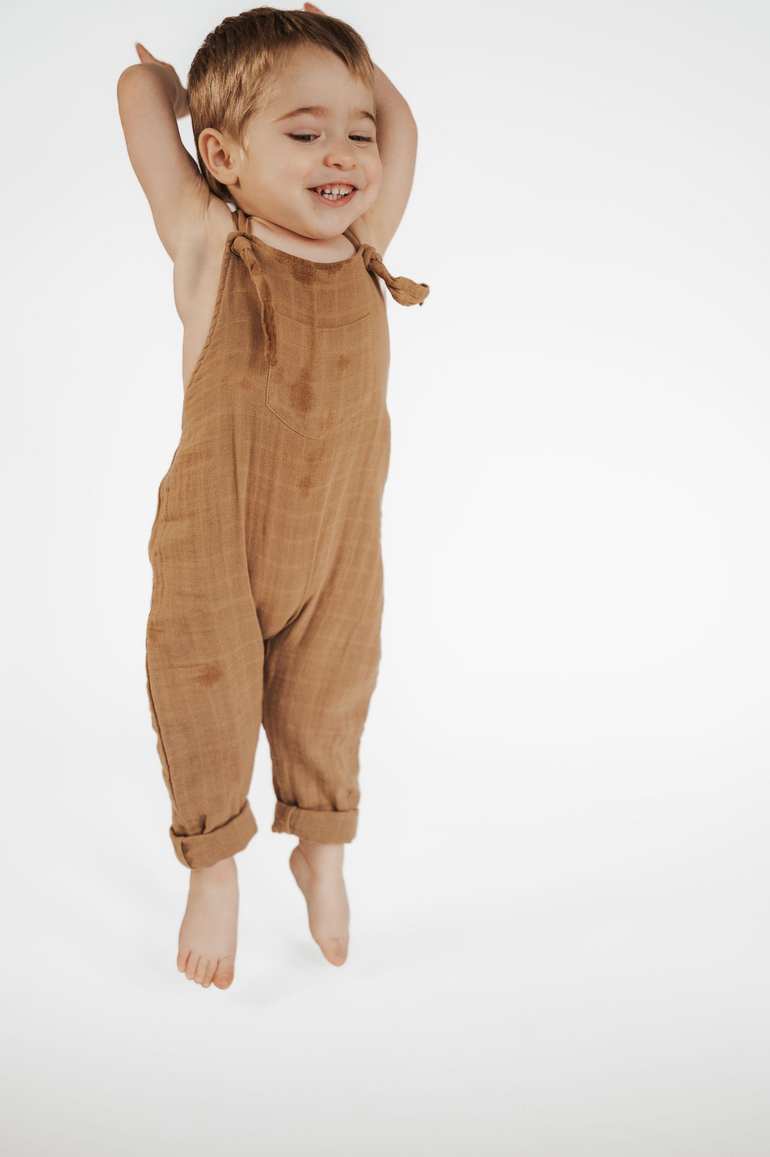 Joyful child wearing brown linen overalls jumps playfully with arms raised against white background.