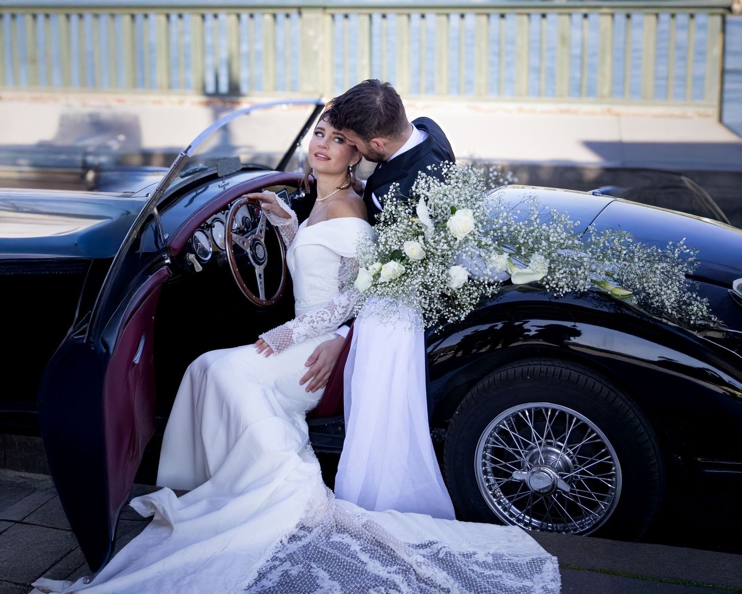 A romantic couple in wedding attire pose next to a classic vintage black car with delicate white flowers.