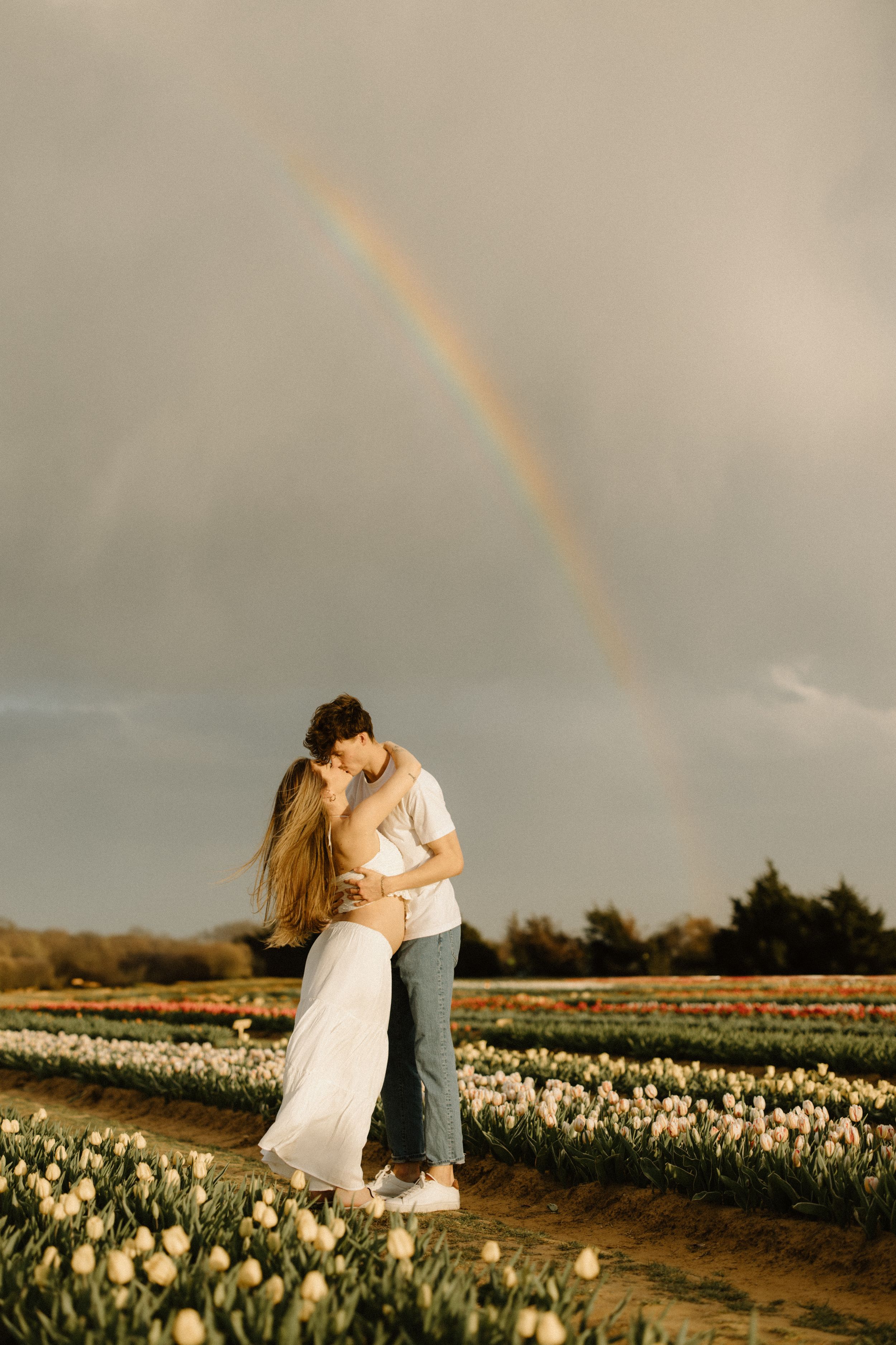 A couple embraces in a field of white tulips at sunset with a rainbow arching through the moody sky.