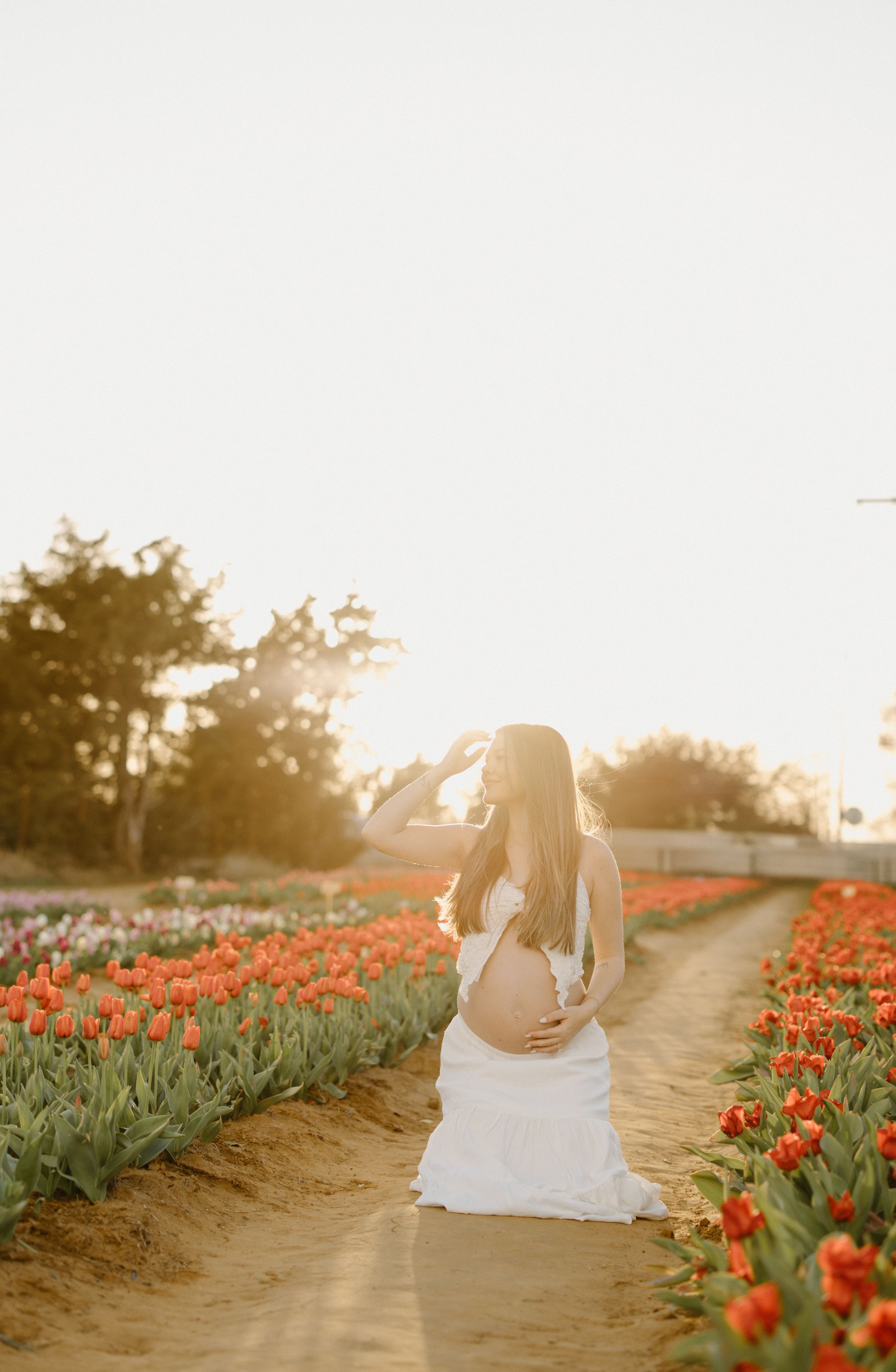 A person in a white dress poses in a tulip field at sunset with vibrant orange flowers lining a dirt path.