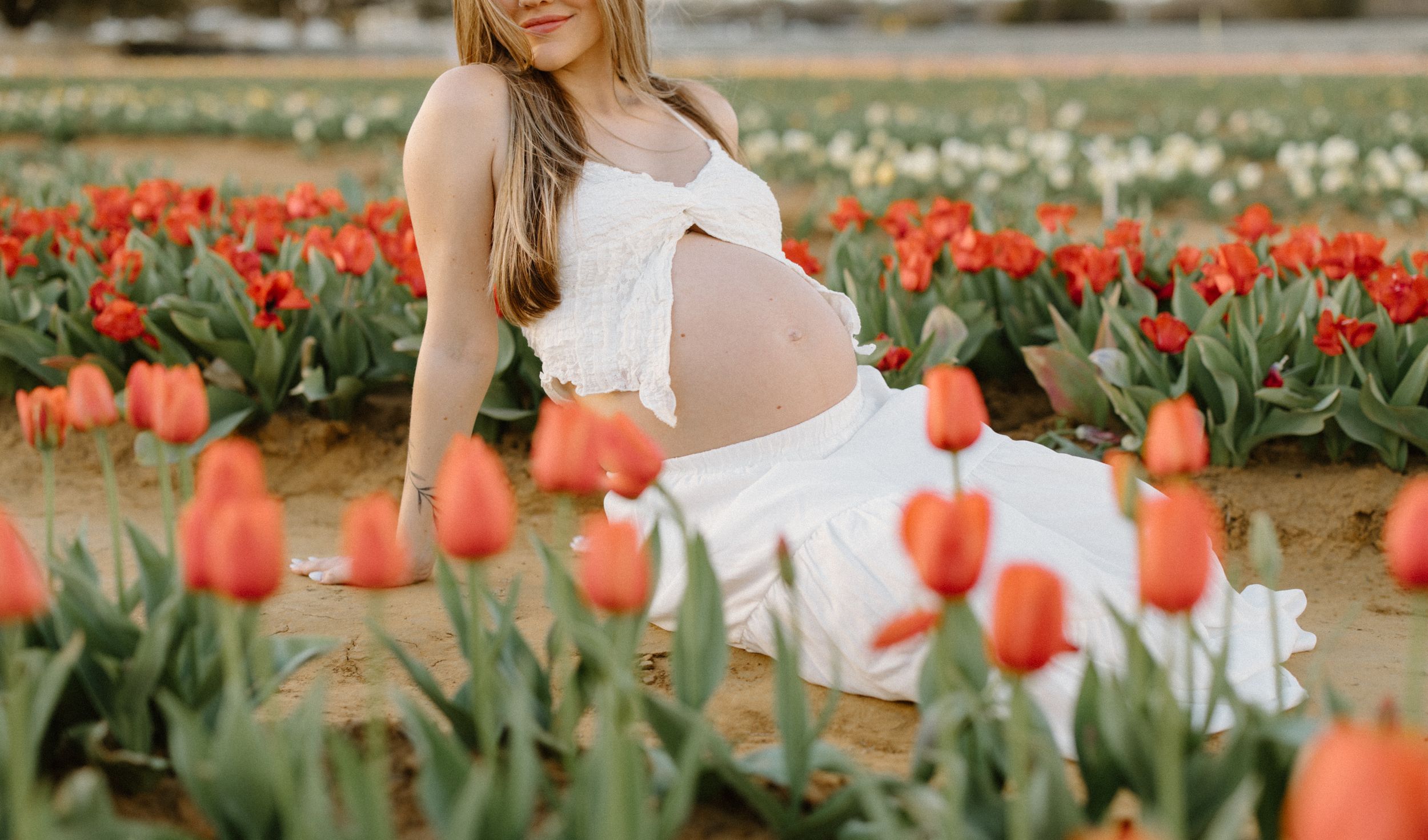 Person in white dress poses among vibrant coral-colored tulips in a blooming spring garden field.