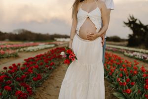 Expecting mother in flowing white dress holds red flowers while standing in vibrant tulip field at sunset.