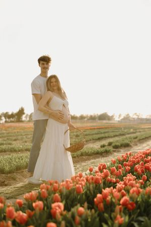 A couple stands together in a field of orange tulips at sunset, with the pregnant woman holding a woven basket.