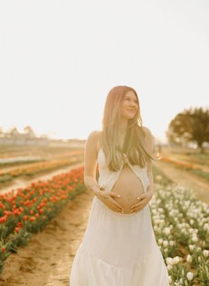 A maternity photo captures an expectant mother in a white dress standing among tulip fields at golden hour.