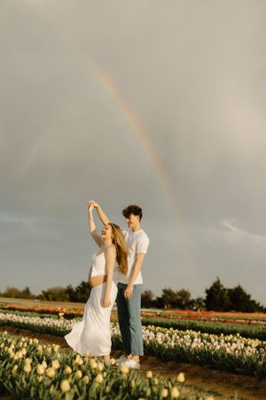 A couple dances in a tulip field at sunset with a rainbow arching across the cloudy sky in the background.